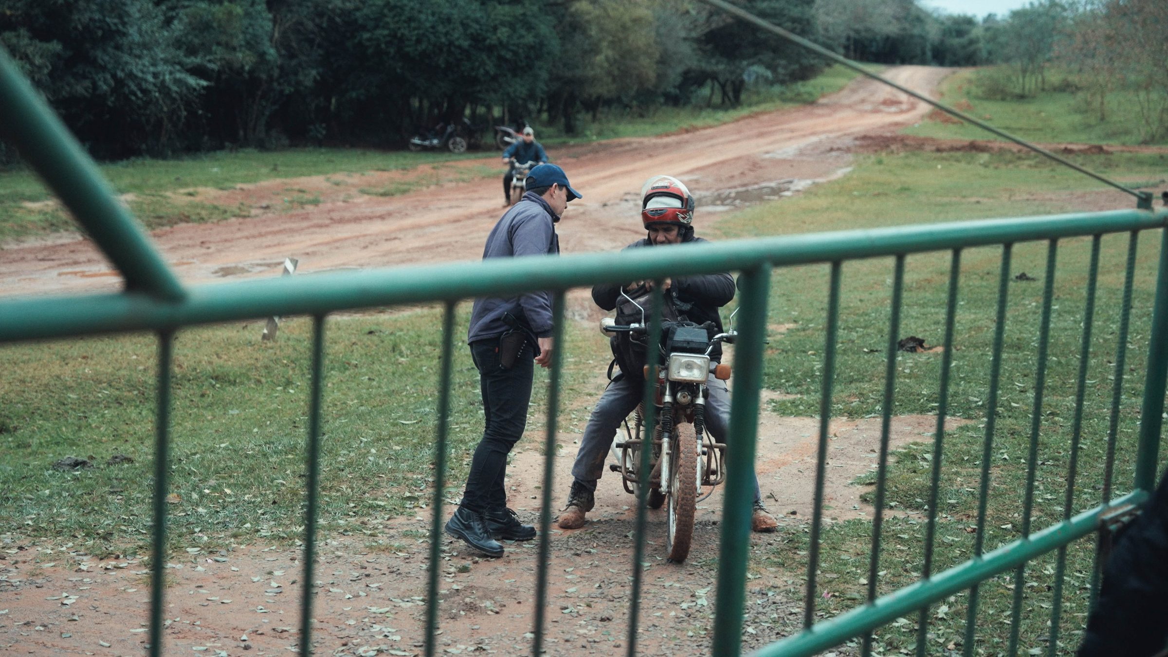 Szene aus Im Umkreis des Paradieses: Zwei Männer an einem grünen Zaun, einer sitzt auf einem Motorrad